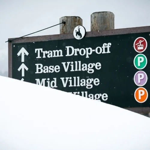 A directional parking sign behind a pile of snow in Teton Village, WY.
